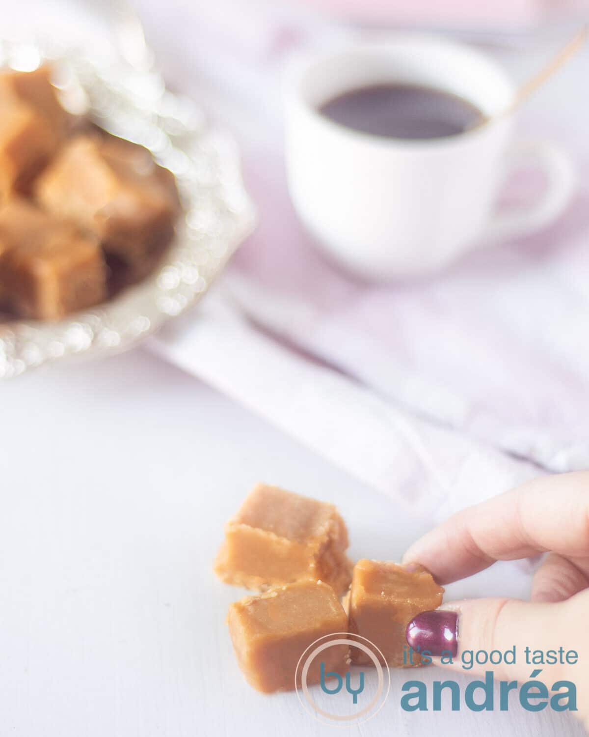 Een hand die een blokje fudge pakt. Een schaaltje met fudge op de achtergrond met een kopje koffie