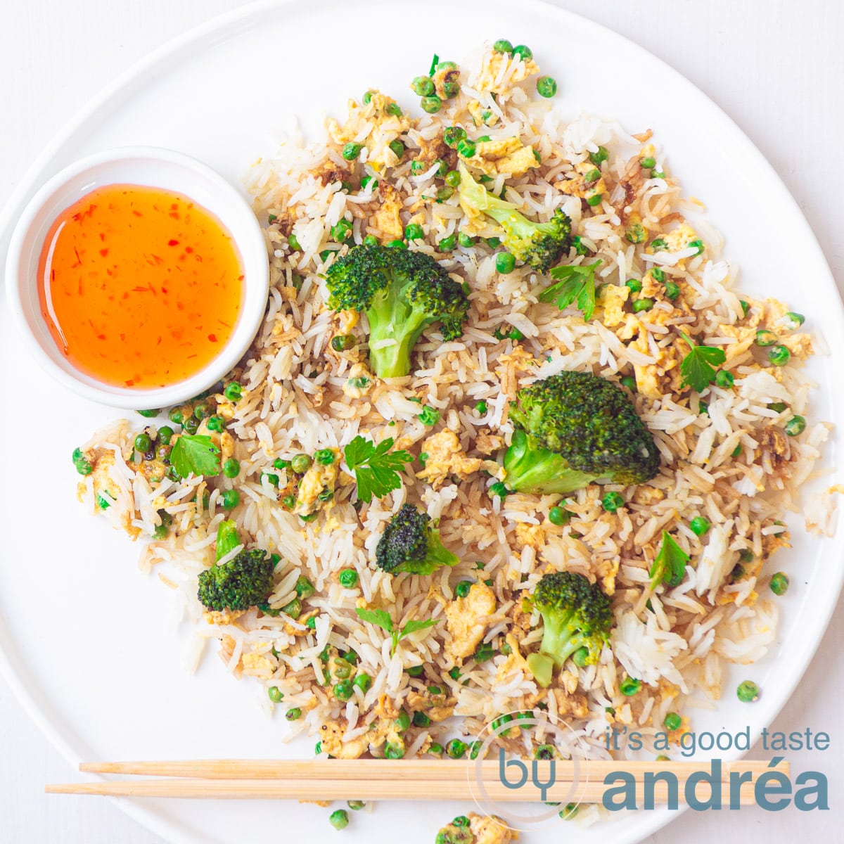Een vierkante foto van een wit bord met gebakken rijst, broccoli, erwtjes en ei. Aan de linkerzijde een kommetje met chilisaus. Onderin twee chopsticks.