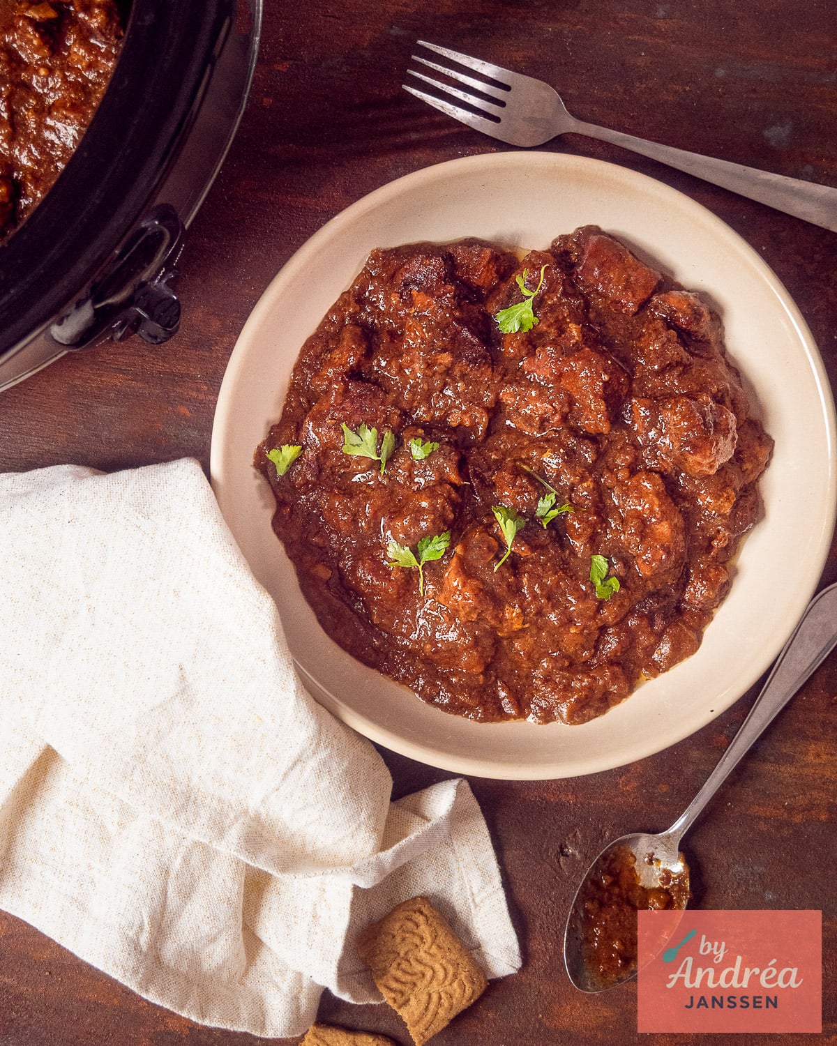 Bovenaanzicht van een bord stoofvlees met peterselie naast de slowcooker, met speculaasjes op een lichte doek.