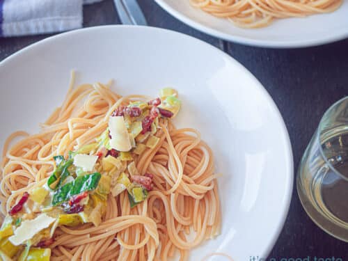 Een vierkante foto met een wit bord met daarop spaghetti, mascarpone, spekjes en gebakken prei op een donkere ondergrond. In de achtergrond een tweede bord.