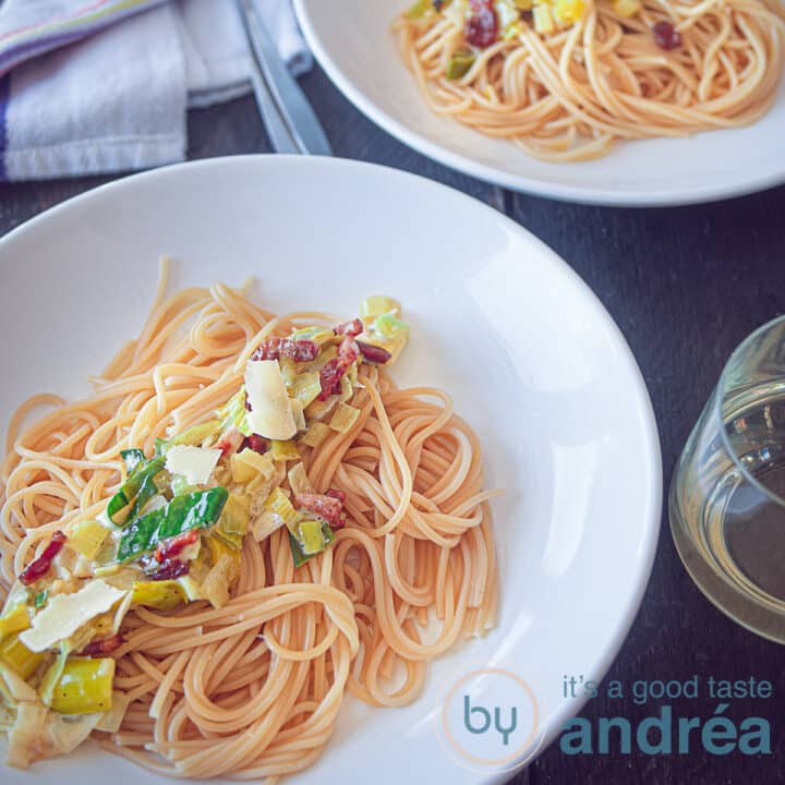 Een vierkante foto met een wit bord met daarop spaghetti, mascarpone, spekjes en gebakken prei op een donkere ondergrond. In de achtergrond een tweede bord.