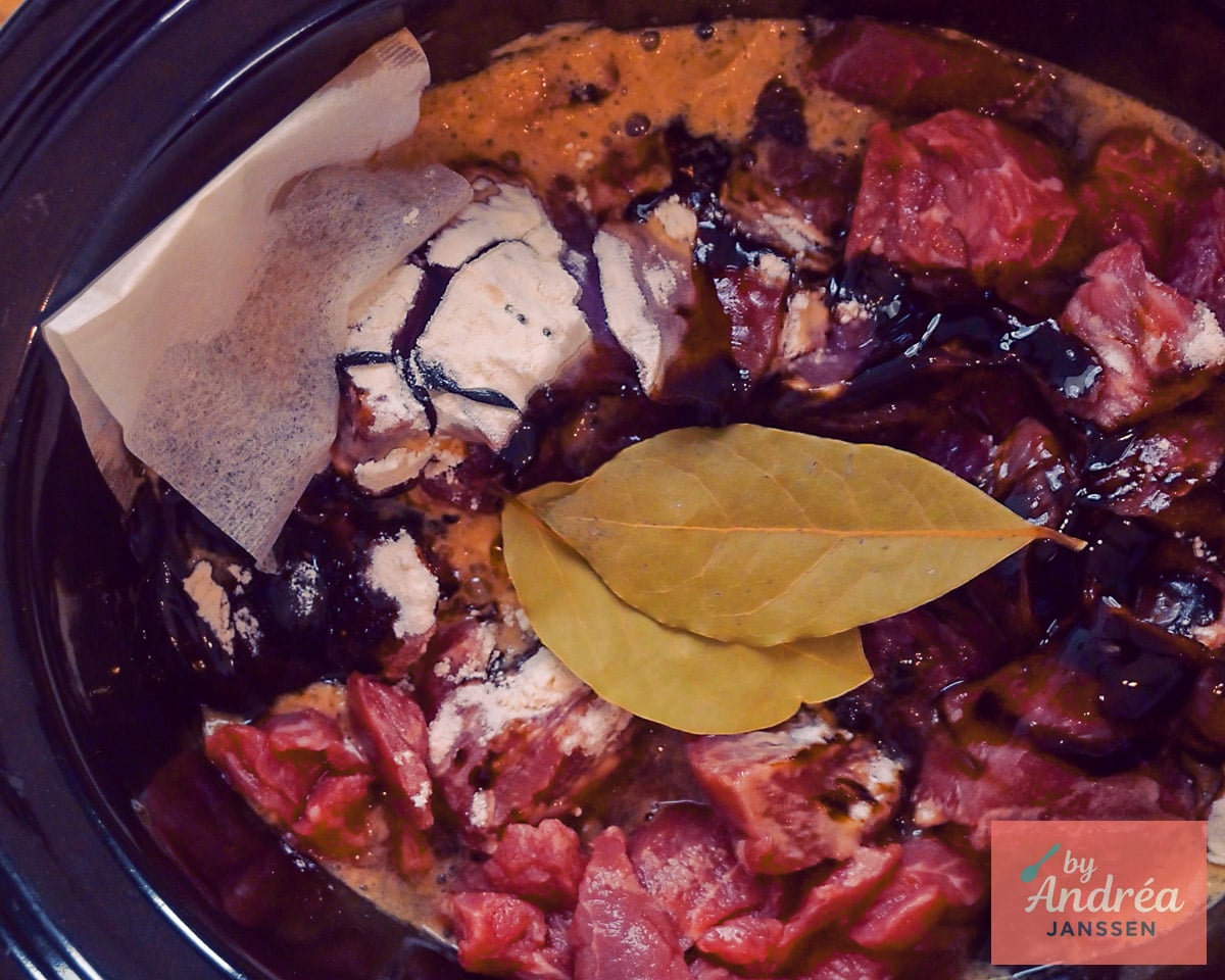 Rauwe rundvleesblokjes met bloem en donkere appelstroop in de slowcooker, met laurierblaadjes en een kruidenzakje.