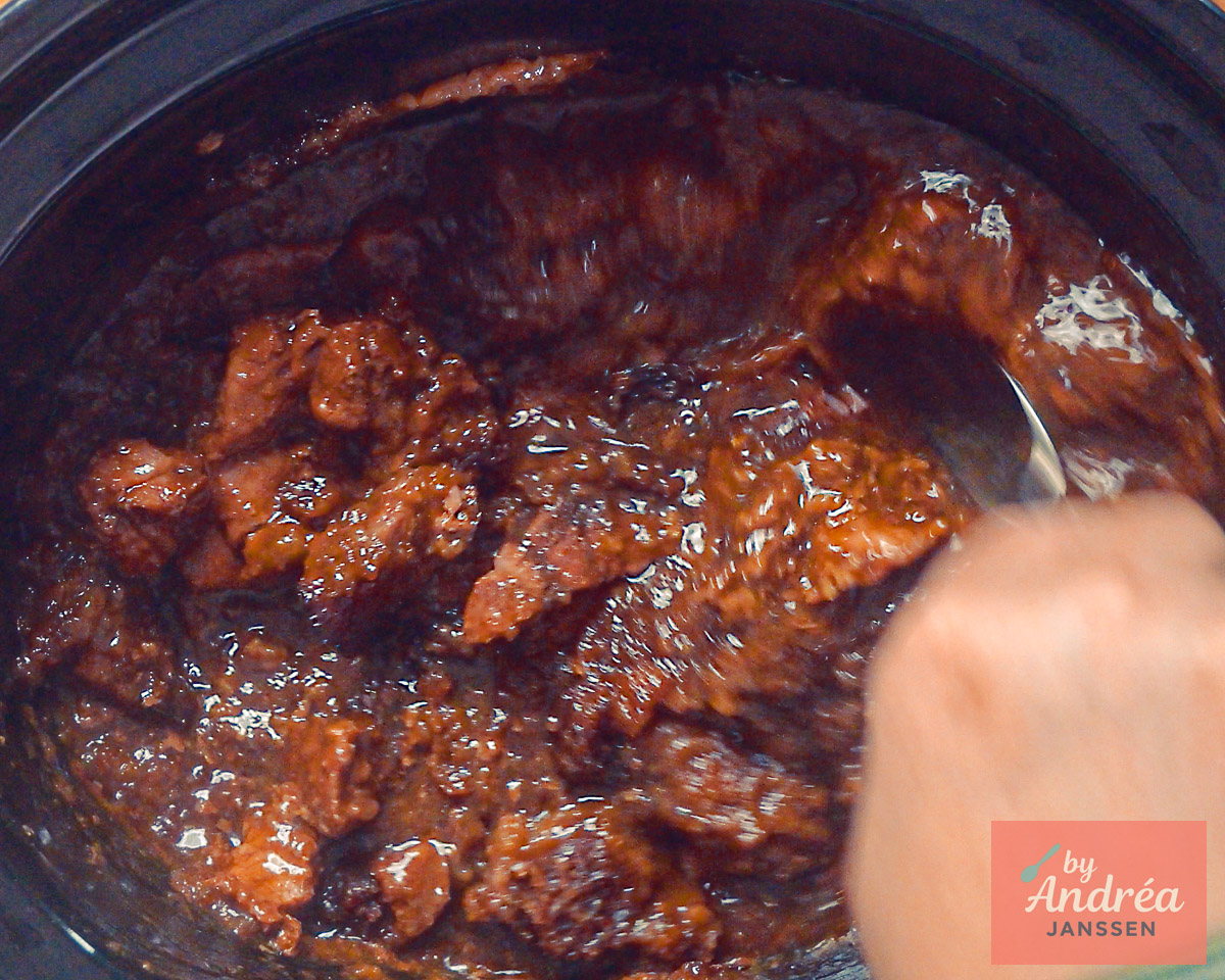 Een hand roert met een lepel door het gare stoofvlees met donkerbruine saus in de slowcooker.