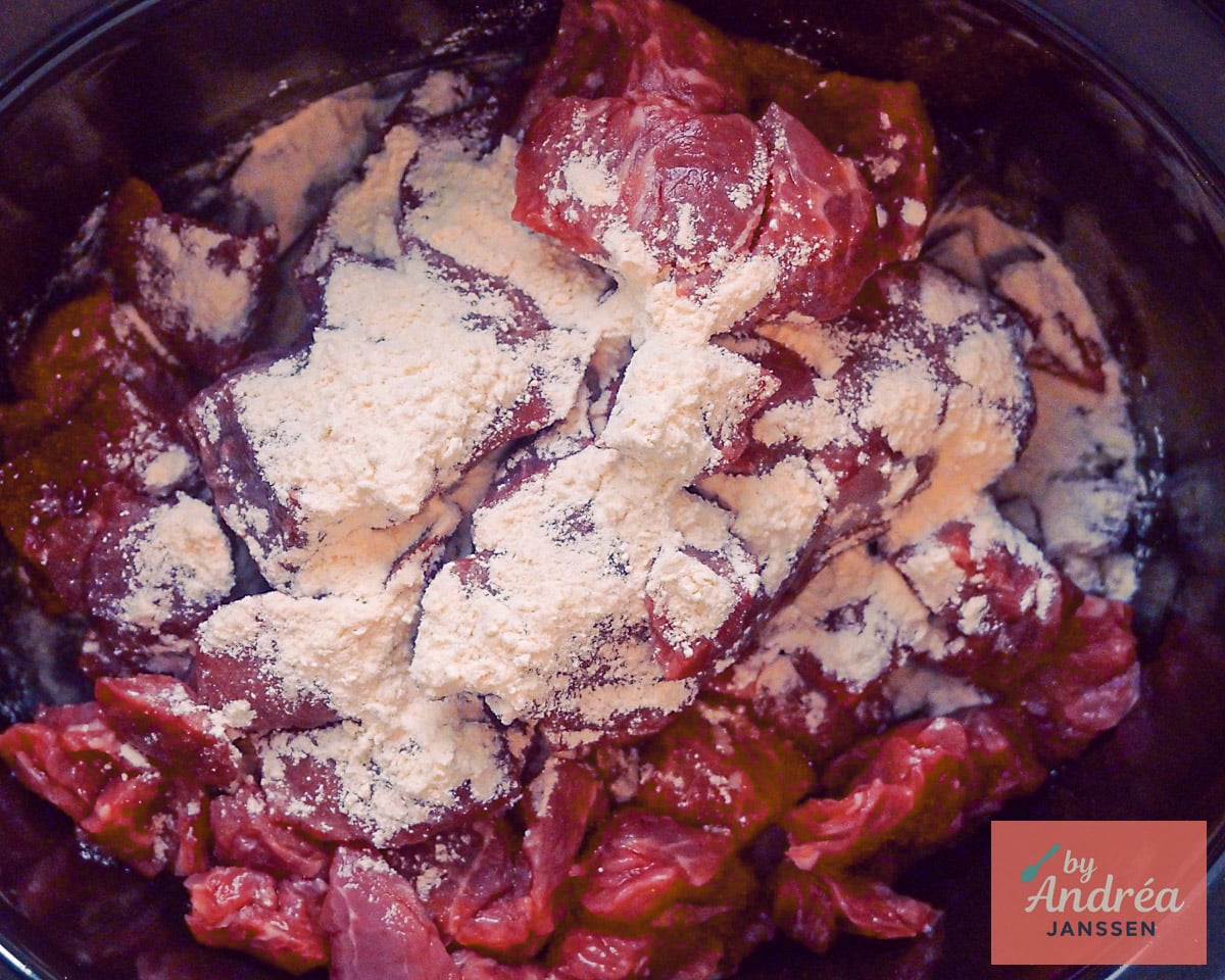 Rauwe blokjes rundvlees bestrooid met bloem in de zwarte binnenpan van de slowcooker.