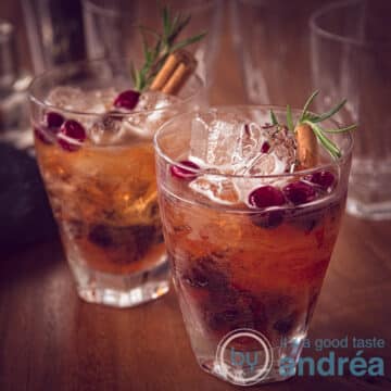 A square photo with two glasses filled with a Prosecco cranberry cocktail diagonally behind each other. With ice cream, cinnamon and rosemary. A wooden tabletop