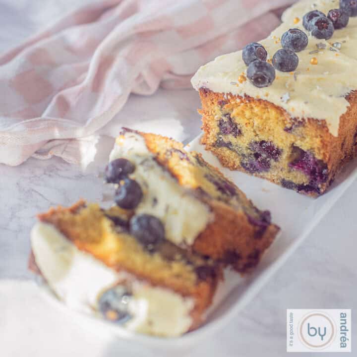 Een vierkante foto met een deel van een cake met blauwe bessen en vanillefrosting. Er zijn twee plakjes afgesneden.