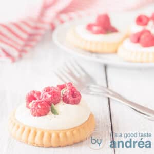 Een vierkante foto van een viertal frambozen gebakjes, één op de voorgrond op een houten witte ondergrond. Twee vorken liggen klaar. Een wit rood gestreepte theedoek in de achtergrond