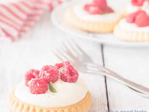 Een vierkante foto van een viertal frambozen gebakjes, één op de voorgrond op een houten witte ondergrond. Twee vorken liggen klaar. Een wit rood gestreepte theedoek in de achtergrond