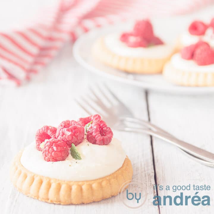 Een vierkante foto van een viertal frambozen gebakjes, één op de voorgrond op een houten witte ondergrond. Twee vorken liggen klaar. Een wit rood gestreepte theedoek in de achtergrond