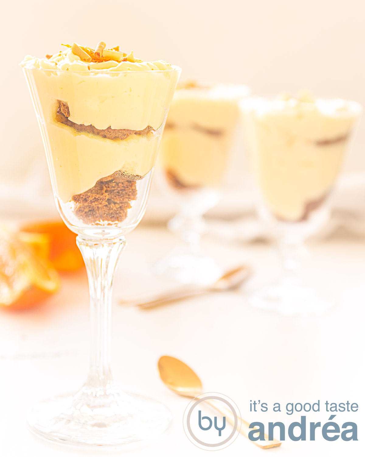 Drie glazen gevuld met sinaasappelpudding en bastognekoekjes. Op de achtergrond sinaasappel en een witte ondergrond.