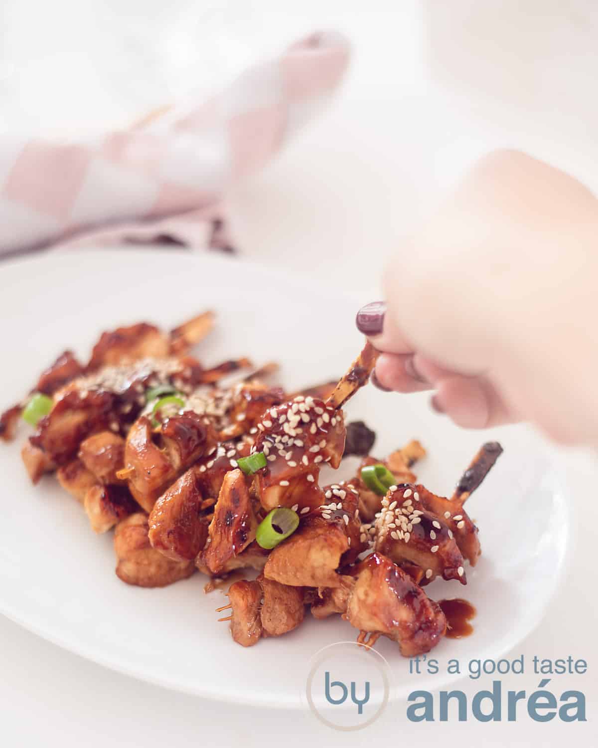 Een witte serveerschaal met een berg yakitori spiesjes. Een hand pakt een stokje van de schaal. Een witte ondergrond