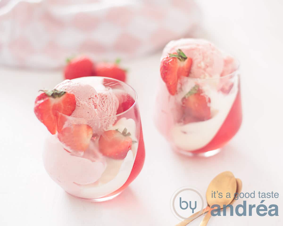 Een breedtefoto met twee glazen gevuld met aardbeien jelly, ijs, verse aardbeien en slagroom. Een witte ondergrond. In de voorgrond twee gouden dessertlepels.