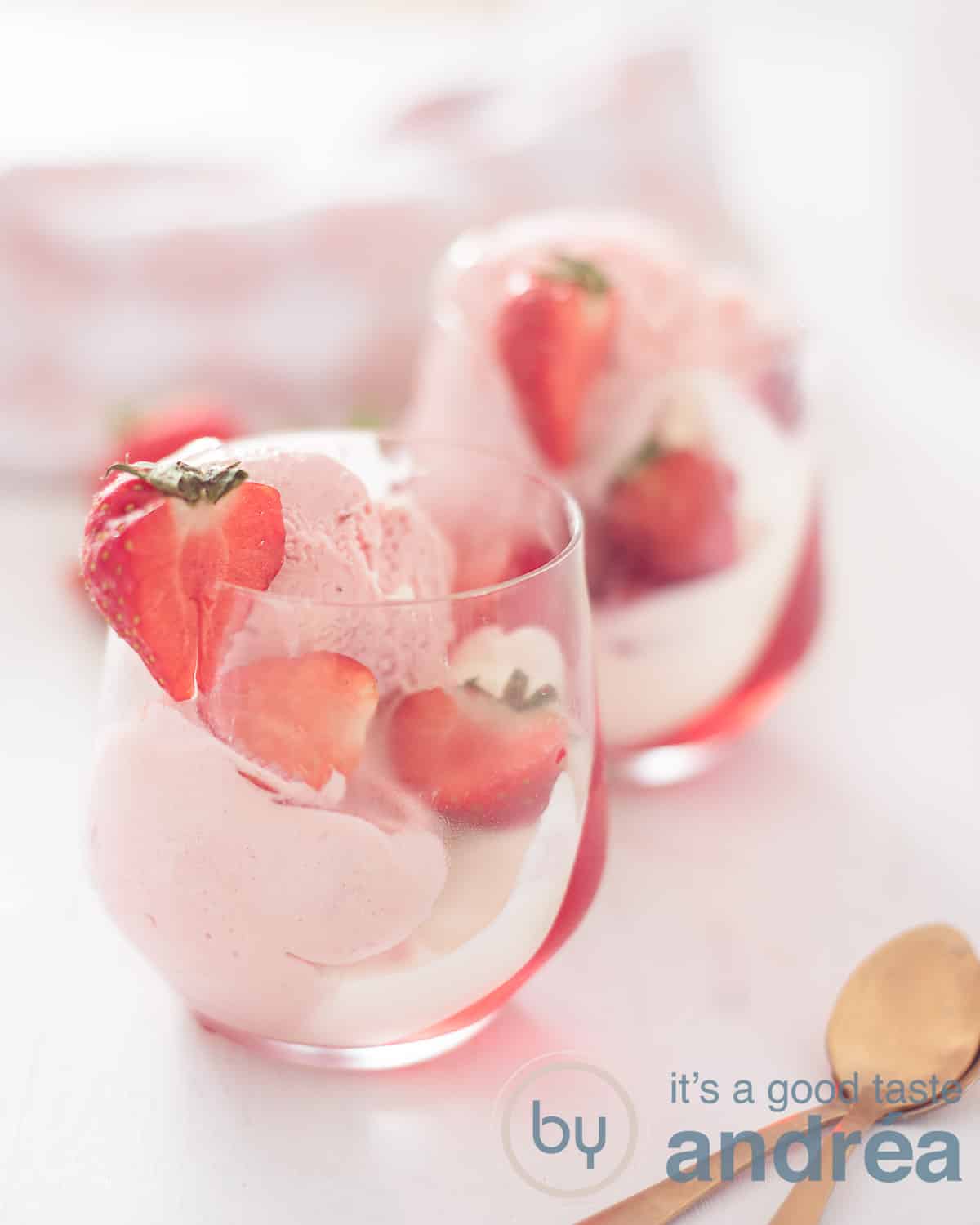 Een hoogtefoto met twee glazen gevuld met een aardbeienpudding, ijs, verse aardbeien en slagroom op een witte ondergrond.