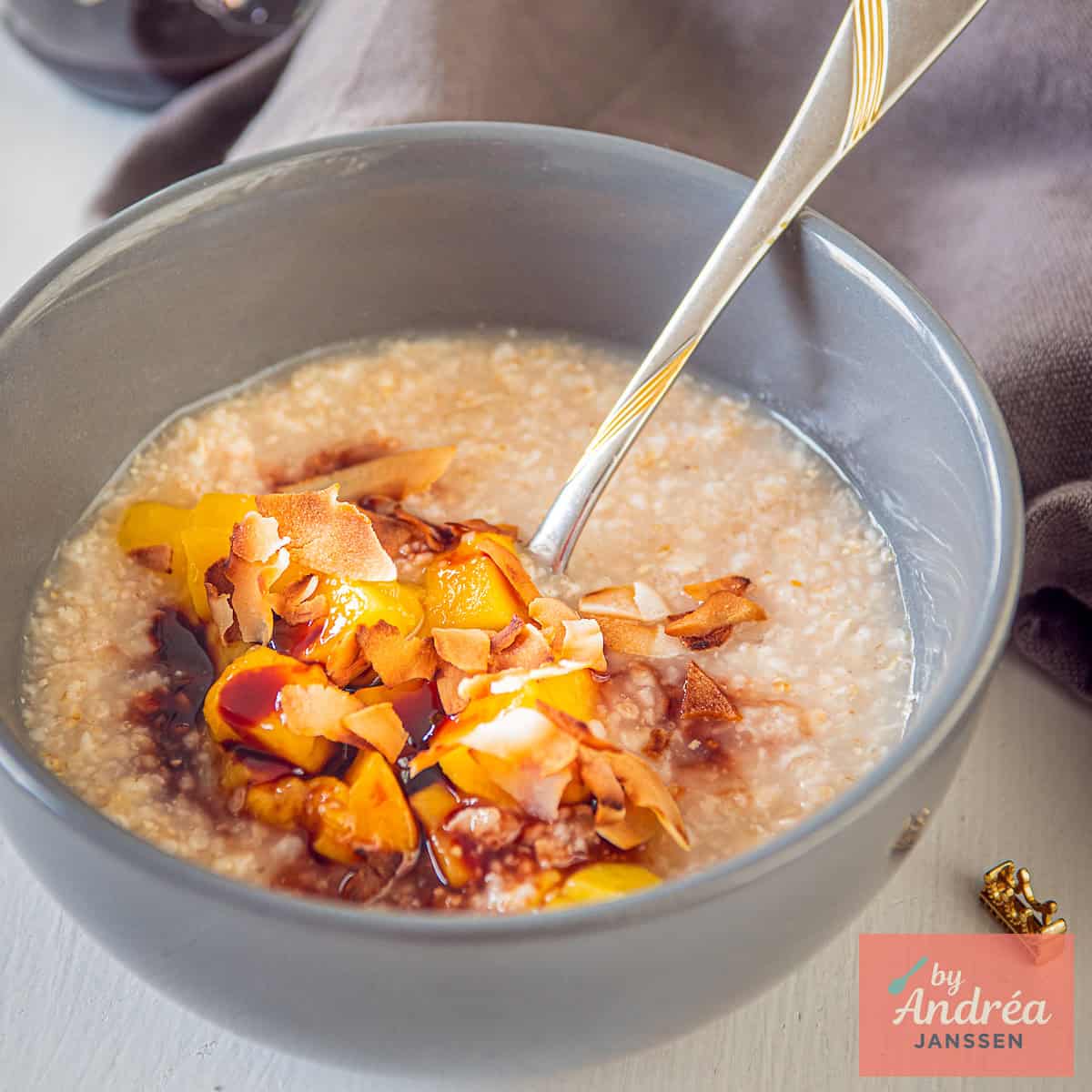 Een vierkante foto met een grijze kom met havermout, mango, dadelstroop en kokos