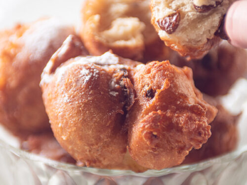 Een glazen schaal met oliebollen. Een hand pakt een oliebol waar een hap uit is genomen. De luchtige structuur is goed te zien.