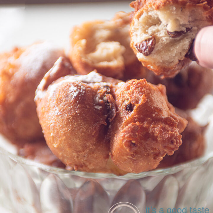 Een glazen schaal met oliebollen. Een hand pakt een oliebol waar een hap uit is genomen. De luchtige structuur is goed te zien.