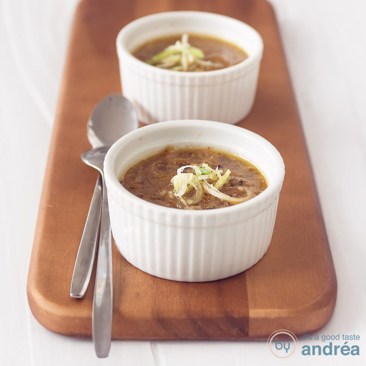 A square photo with two bowls filled with leek soup with cabbage. Two spoons next to it on a wooden board. A white background.
