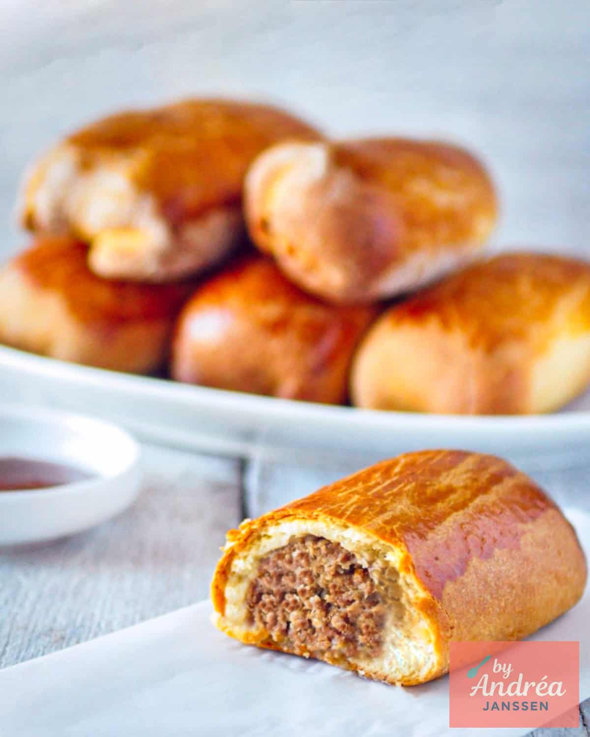 Een bord met Brabantse worstenbroodjes. Vooraan een doormidden gesneden worstenbroodje, vulling zichtbaar.