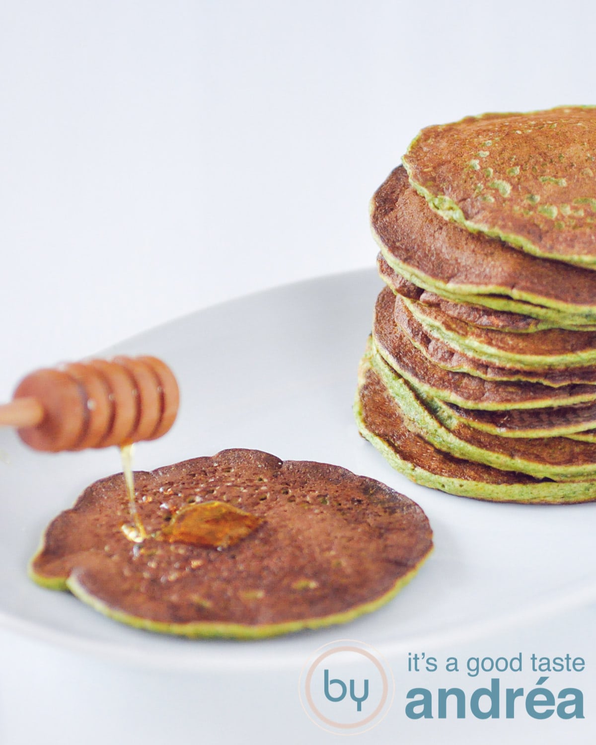 Een wit bord met aan de linkerkant een gorzen pancake met honing drizzle en aan de rechterkant een stapel groene pancakes