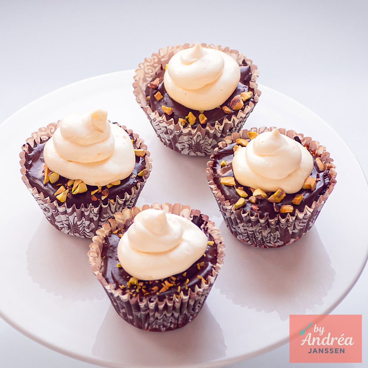 Een vierkante foto met vier cupcakes op een wit taart plateau met ganache, witte chocolade boter crème en pistache.