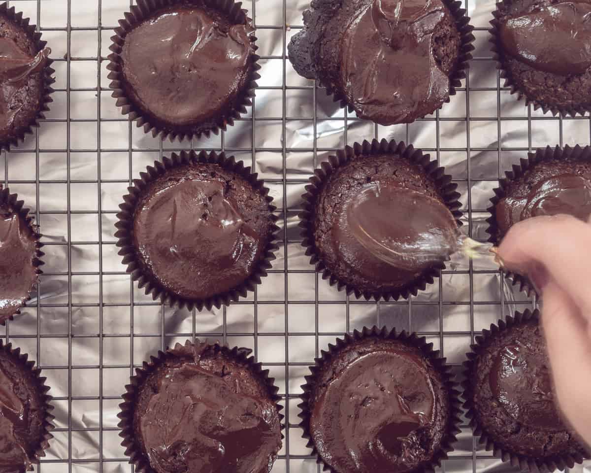 Chocolade cupcakes op een rooster. Met een lepel wordt er ganache overheen gesmeerd.