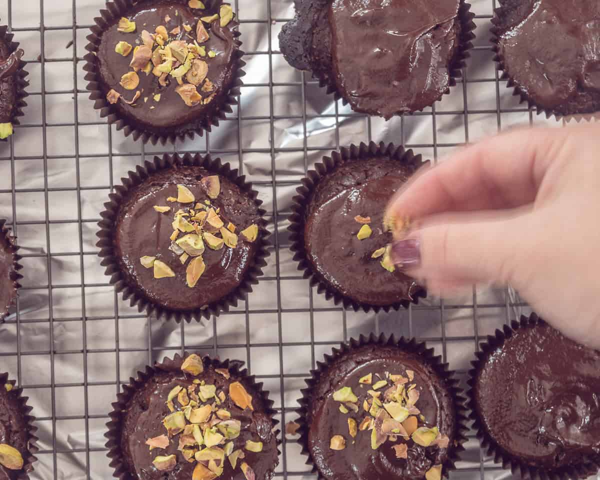Cupcakes op een rooster. Een hand strooit er wat gehakte pistachenoten overheen.