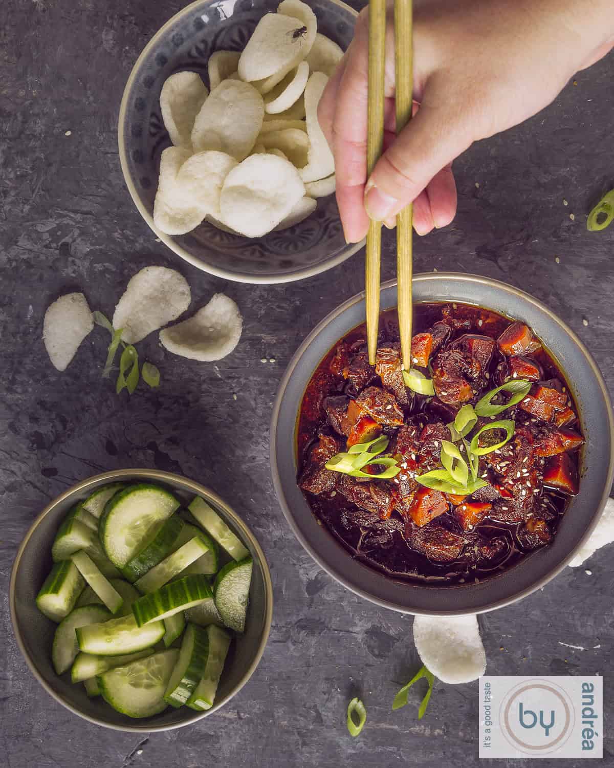 Een foto met drie grijze kommen gevuld met Mongools rundvlees, kroepoek en komkommersalade. Twee eetstokjes die wat vlees pakken