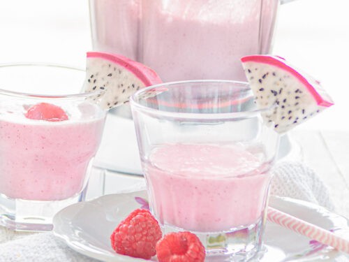 Een vierkante foto met een wit bord daarop een glas gevuld met dragon fruit smoothie met frambozen en een schijfje drakenfruit. Een witte ondergrond