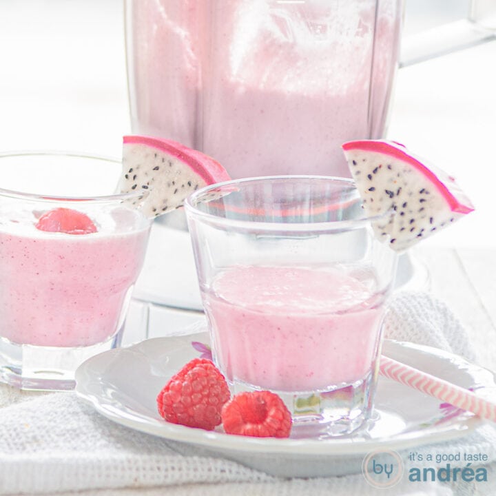 Een vierkante foto met een wit bord daarop een glas gevuld met dragon fruit smoothie met frambozen en een schijfje drakenfruit. Een witte ondergrond