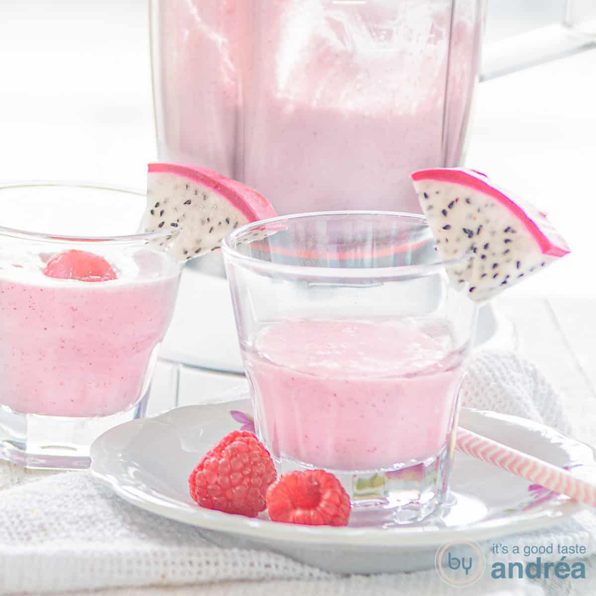 Een vierkante foto met een wit bord daarop een glas gevuld met dragon fruit smoothie met frambozen en een schijfje drakenfruit. Een witte ondergrond