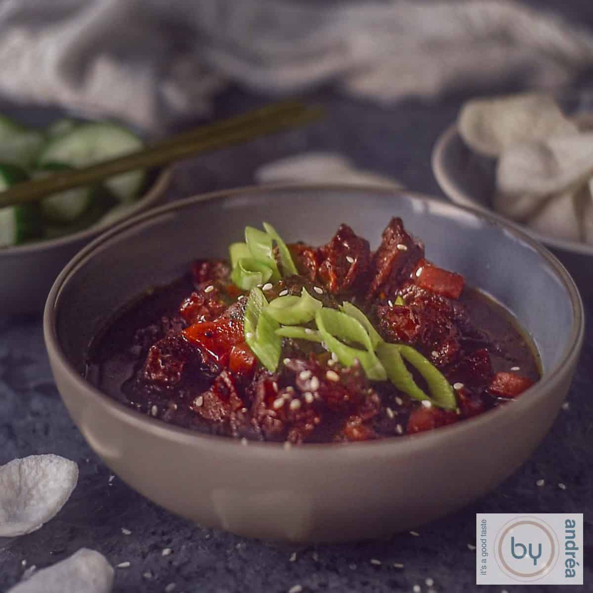 Een vierkante foto met een grijze kom gevuld met Mongools rundvlees uit de slowcooker, met lente-ui als garnering en wat kroepoek.