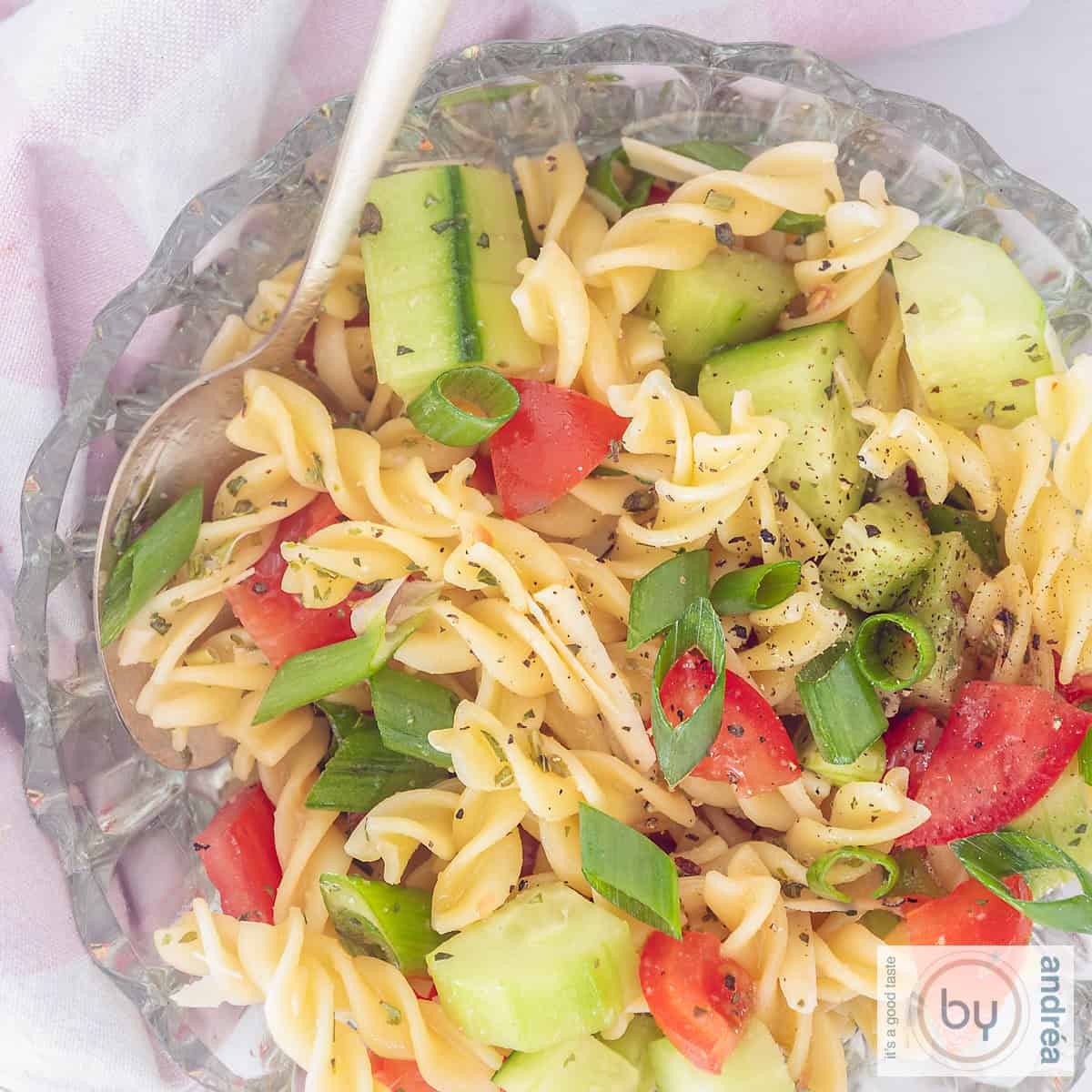 Een vierkante afbeelding, met de bovenkant naar beneden, van een glazen kom gevuld met fusillisalade met komkommer, lente-uitjes, tomaten en dressing.