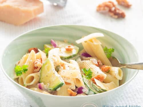 Een vierkante foto met een groen schaaltje met penne, walnoten, courgette en Parmezaanse kaas op een witte ondergrond. Een stuk parmezaanse kaas en walnoten in de achtergrond