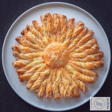 Een vierkante foto van een donkere ondergrond met een wit bord met een tarte soleil van bladerdeeg en Boursin.