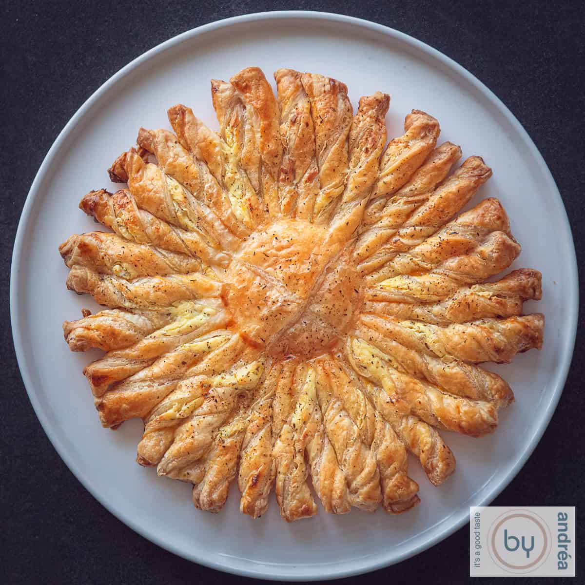 Een vierkante foto van een donkere ondergrond met een wit bord met een tarte soleil van bladerdeeg en Boursin.