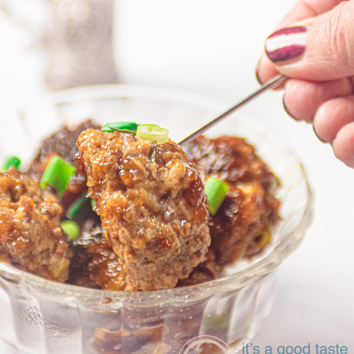 Een glazen schaaltje met gehaktballetjes met een teriyaki glaze en lente-ui. Een zilveren prikker pakt een balletje. In de achtergrond een zilveren bakje met meer prikkers en een opscheplepel