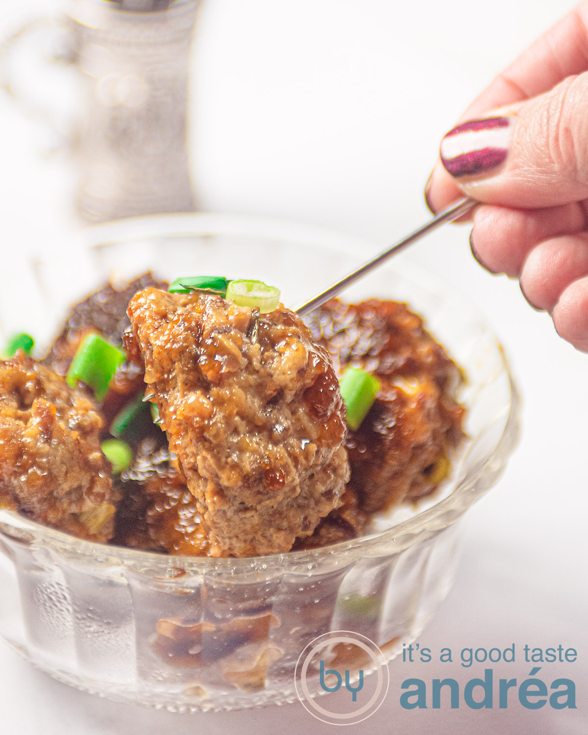 Een glazen schaaltje met gehaktballetjes met een teriyaki glaze en lente-ui. Een zilveren prikker pakt een balletje. In de achtergrond een zilveren bakje met meer prikkers en een opscheplepel