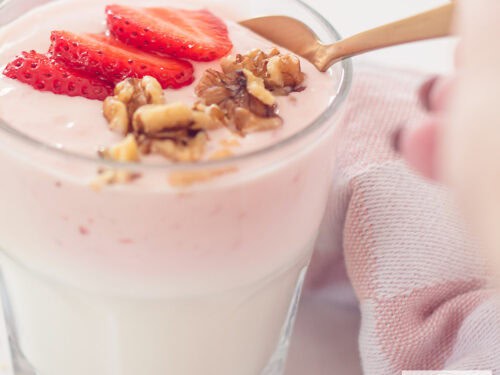 Een vierkante foto van een glas gevuld met zelfgemaakte aardbeien yoghurt, walnoten en verse aardbeien. Een gouden lepel neemt een schep.
