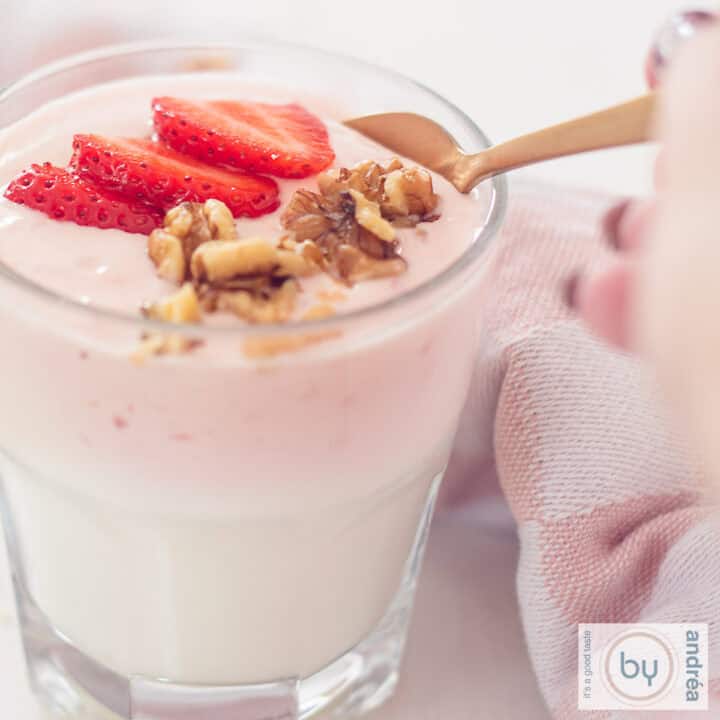 Een vierkante foto van een glas gevuld met zelfgemaakte aardbeien yoghurt, walnoten en verse aardbeien. Een gouden lepel neemt een schep.
