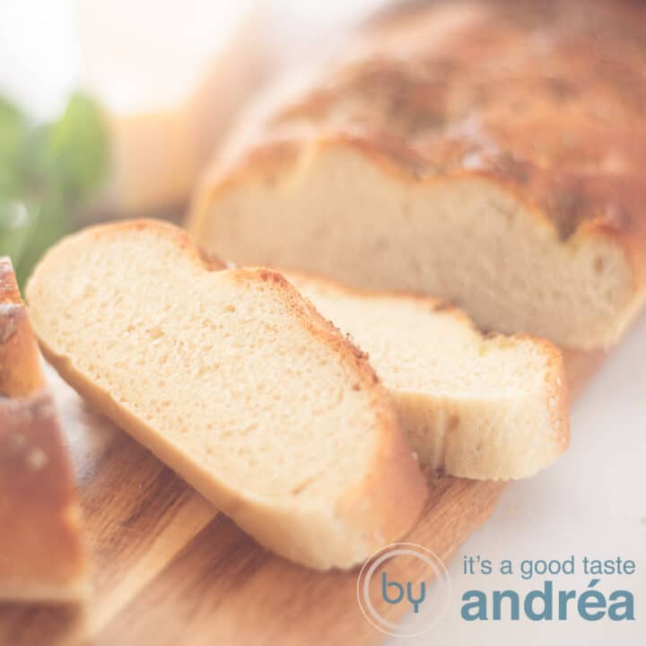 Een vierkante foto met een deel van een Italiaanse focaccia met een topping van basilicum en Parmezaanse kaas. Twee sneden eraf gesneden. Alles op een houten plank op een witte odnergrond