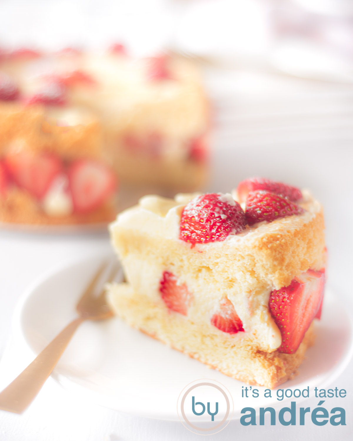 Een stuk aardbeien chiffon cake op een wit bord met een gouden vork. In de achtergrond de rest van de taart