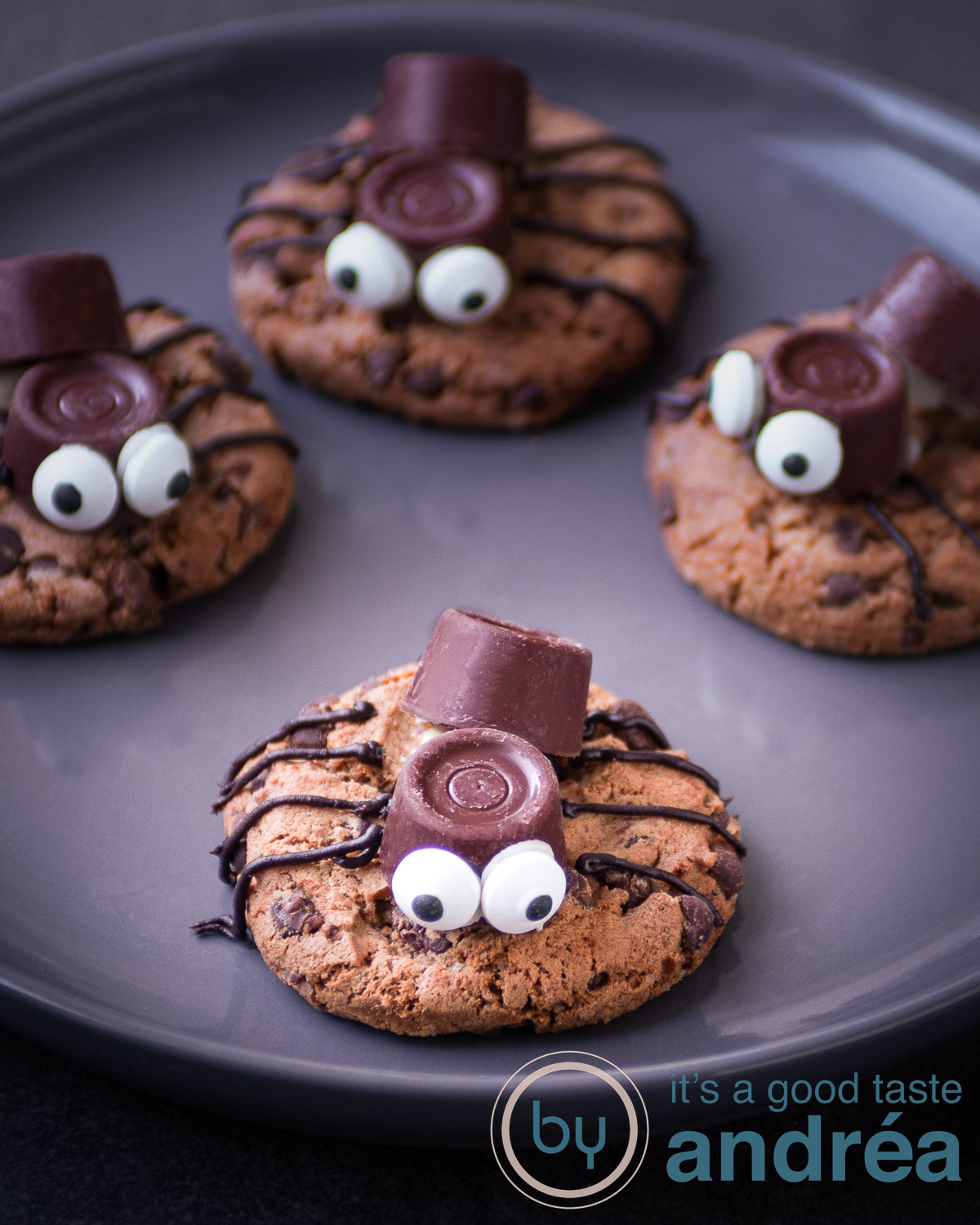 Een koekje met een spinnenrug, twee oogjes en chocolade pootjes omringt met nog drie koekjes op een grijs bord