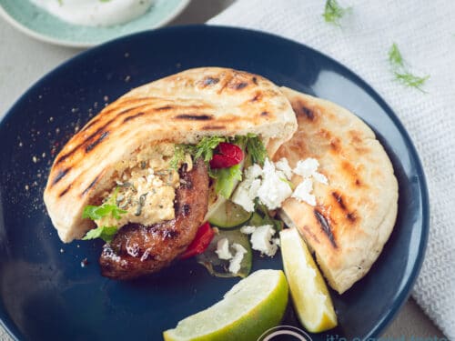 een vierkante foto met een blauw bord met daarop een pita broodje met runderhamburger en gegrilde groenten