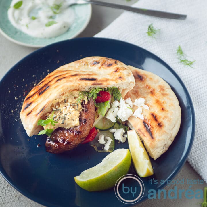 een vierkante foto met een blauw bord met daarop een pita broodje met runderhamburger en gegrilde groenten