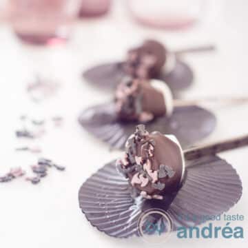 A square photo with three marshmallow pops in a row, dipped in chocolate and sprinkles on a black cupcake round. Sprinkles on the left. A white background.