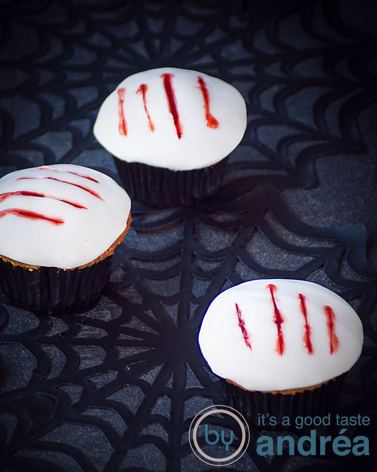 Drie cupcakes met een witte toplaag en vier klauw afdrukken gevuld met aardbeien bloed. Ze zijn geplaatst op een spinnenweb.