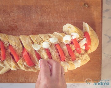 Een houten plank met een stokbrood gevuld met tomaat waartussen de plakjes mozzarella wordt gestopt