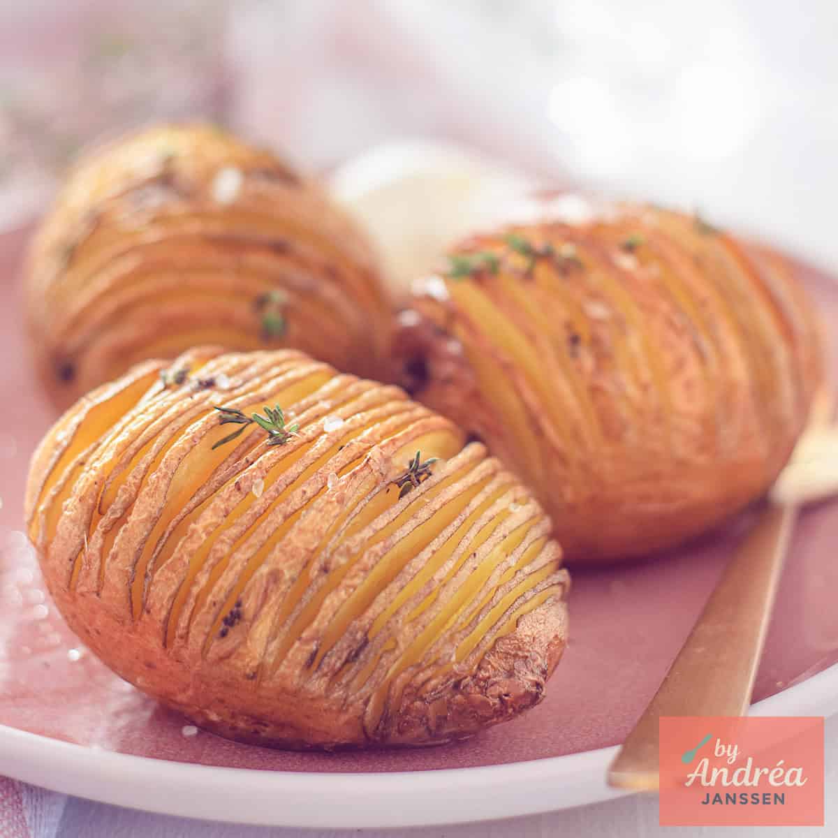 Een vierkante foto met drie hasselback aardappelen en crème fraîche op een roze bord.