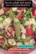 Een deel van een roze schaal met een salade met rucola, tomaat, peer, appel en pistachenoten. Een tekst: Rucola salade met appel, peer en pistachenoten