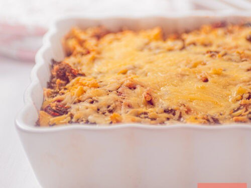 Een vierkante foto met een deel van een witte ovenschaal gevuld met een gratin van boerenkoolstamppot, kaas, en tomaat